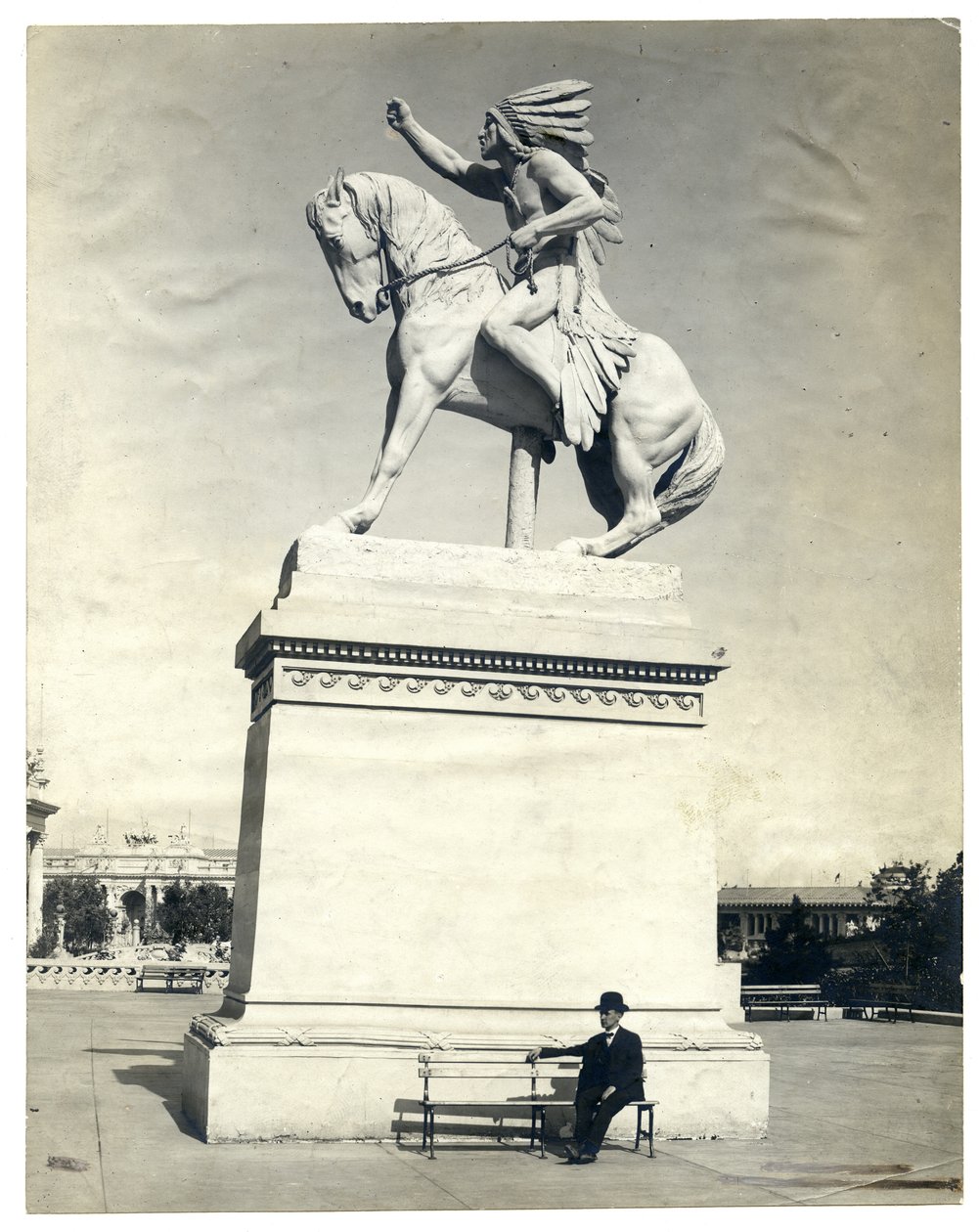 Büyük bir kaidenin üzerinde bir at üzerinde oturan bir Kızılderili heykelini gösteren dikey, sepya fotoğraf. Kaide tabanında bir bankta oturan koyu renk takım elbiseli ve şapkalı bir adam var. by Unknown